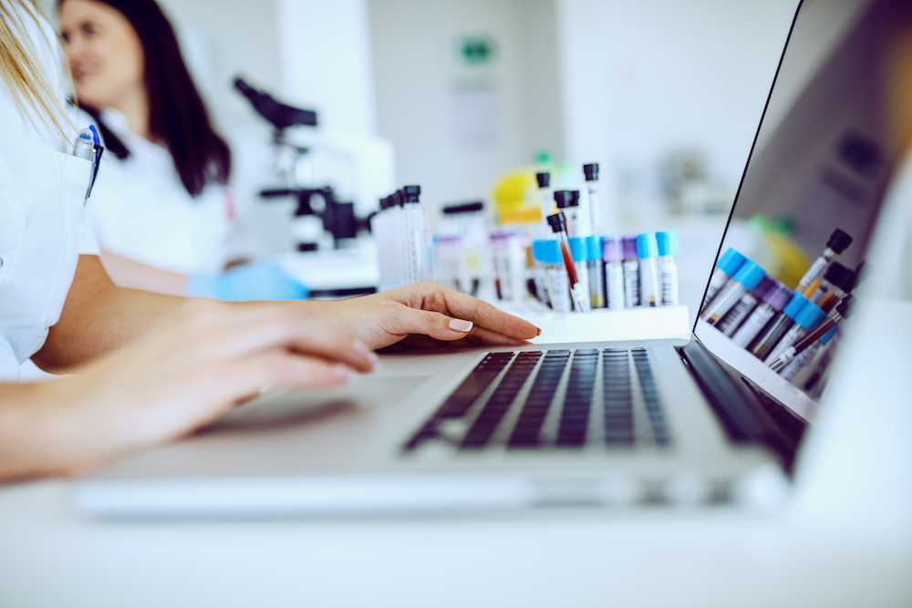 Close up of female lab assistant in white uniform sitting in lab and using laptop for data entry. In background is her colleague working.